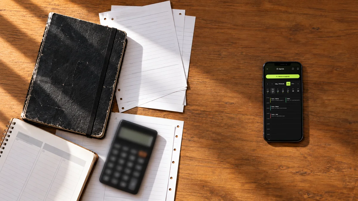 Caderno de treinos gasto, fichas soltas, agenda de papel e calculadora de um lado da mesa; do outro, só o celular aberto na agenda da Welltrainer