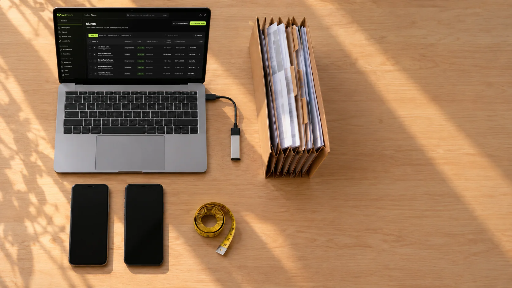 Vista de cima de uma mesa com notebook aberto na lista de alunos da Welltrainer, dois celulares, pendrive e uma pasta de fichas em papel