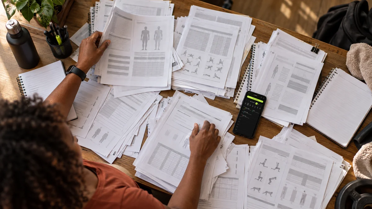 Personal trainer tenta organizar dezenas de fichas de alunos espalhadas pela mesa, com o celular aberto na agenda da Welltrainer no meio da bagunça