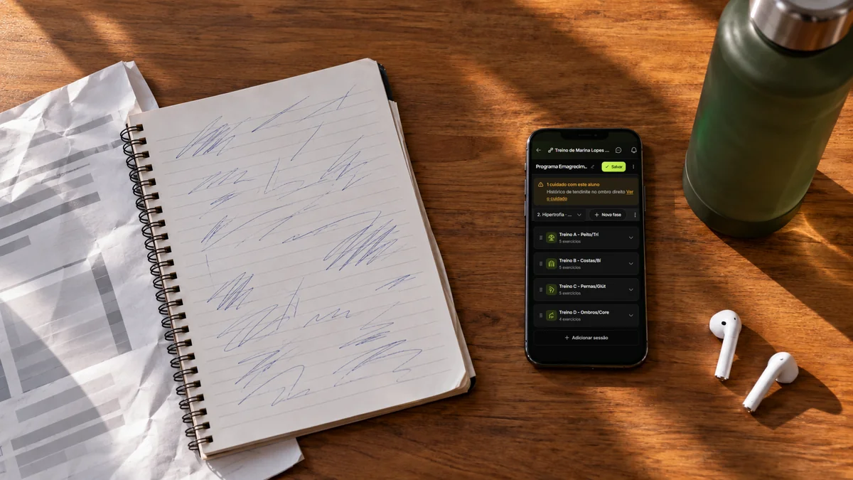 Caderno com treino rabiscado à mão e folha amassada ao lado do celular aberto no programa de treino organizado da Welltrainer